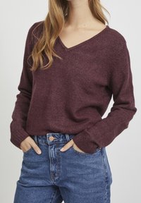 Pull bordeaux à col en V, fabriqué en tissu tricoté texturé, avec des manches longues et des poignets côtelés, associé à un jean bleu taille haute.