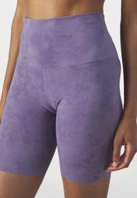 Gros plan d'une personne portant un cuissard de vélo Nike violet taille haute avec une texture subtile et un logo sur la hanche gauche.