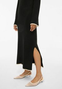 Černé šaty ke kolenům s bočním rozparkem, s dlouhými rukávy a kontrastním krémovým lemováním. Kombinované s béžovými botami na podpatku se špičatou špičkou a páskem za patou.