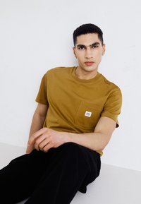 T-shirt in cotone giallo senape con maniche corte, scollatura rotonda e taschino sul petto con un piccolo logo. Pantaloni neri completano l'outfit.