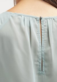 Blusa verde chiaro con dettaglio a keyhole sul retro, texture morbida, collo plissettato e piccolo bottone argentato decorativo sul collo.