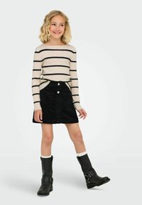 Uśmiechnięta młoda dziewczyna z kręconymi blond włosami, ubrana w kremowy sweter w czarne paski, czarną spódnicę, białe skarpetki i czarne buty, stojąca na białym tle.