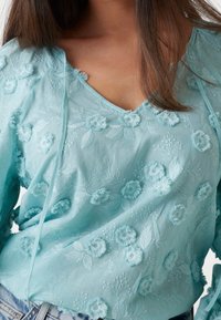 Blouse bleu clair avec broderie florale, décolleté en V et manches longues. Présente des appliqués de fleurs en 3D texturés et des motifs de feuillesintriqués.