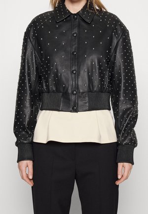 Schwarze, kurz geschnittene Lederjacke mit silbernen Nieten, Knopfleiste vorne, gerippten Bündchen und Bund, getragen über einer weißen Bluse und schwarzen Hosen.