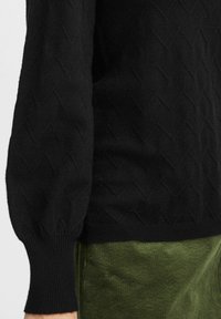 Schwarzer Strickpullover mit einem strukturierten Rautenmuster, puffigen Ärmeln und gerippten Bündchen, kombiniert mit olivgrünen Cordhosen.