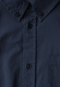 Camisa de botón azul marino con cuello tradicional, botones negros y un bolsillo en el pecho. Tejido suave con una textura fina.