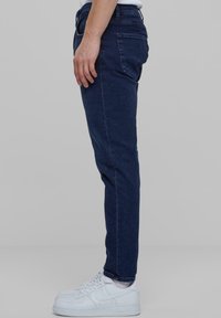 Sötétkék farmernadrág, slim fit szabással, öt zsebes kialakítással, varrott részletekkel és gombos záródással. Fehér sportcipővel viselve.