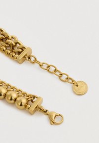 Kuldsest topelt-ahelast käevõru, millel on homaariklamber ja reguleeritavad lingid, ning milles on väike ümmargune ripats, millel on graveering "Liu Jo."