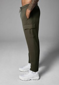 Olivgrüne Jogginghosen mit einem Kordelzugbund und Seitentaschen, ausgestattet mit einer Cargo-Tasche am Oberschenkel. Kombiniert mit weißen Sneakers.