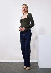Mørkegrønn, teksturert langermet topp med firkantet utringning og volanger, sammen med høyt liv mørkeblå jeans og beige hæler.