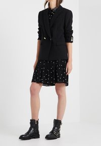 Blazer nero su misura con bottoni dorati, indossato sopra un abito nero con motivi di stelle dorate; abbinato a stivali neri alla caviglia.