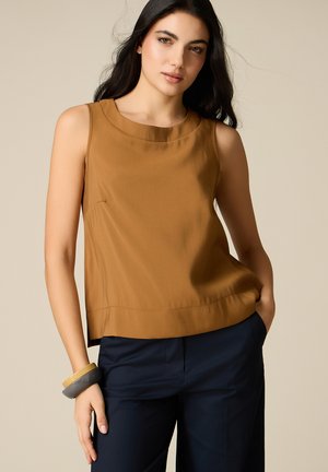 Femme aux longs cheveux foncés portant un haut marron sans manches, un pantalon bleu marine et des bracelets épais, posant avec une main dans la poche.