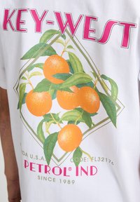 Witte T-shirt met een grafisch ontwerp van sinaasappels en groene bladeren, met bovenaan "KEY-WEST" in het roze en "PETROL IND" in het rood eronder.