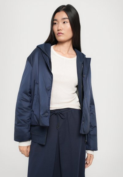 Temno modra pufasta jakna s kapuco, z zadrgo spredaj, gumbnimi poudarki in elastičnimi manšetami. Obuta čez svetlo obarvano dolgimi rokavi.