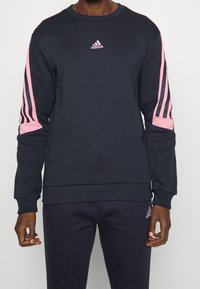 Model wears a navy sweatshirt with pink stripes on the sleeves and a small Adidas logo on the chest, paired with matching navy pants.