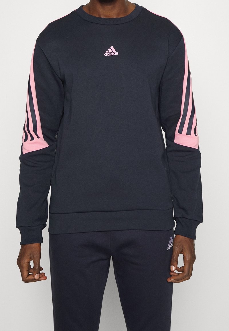 Model wears a navy sweatshirt with pink stripes on the sleeves and a small Adidas logo on the chest, paired with matching navy pants.