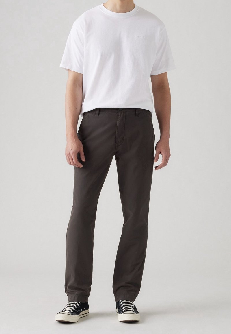 Homme portant un T-shirt blanc à manches courtes, un pantalon marron foncé et des baskets basses noires, debout devant un fond clair uni.