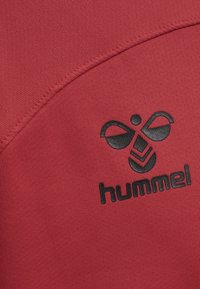 Rød stof med en tekstureret overflade og et sort logo design, der inkluderer buede elementer og ordet "hummel" i fede bogstaver.