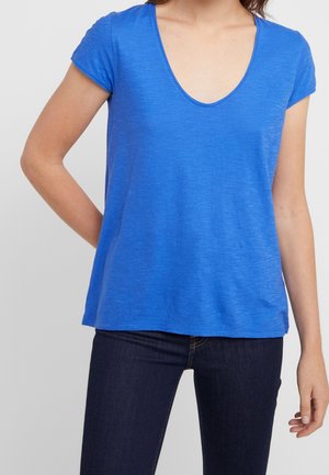 Frau, die ein blaues kurzärmeliges V-Ausschnitt-Oberteil und dunkelblaue Jeans trägt, steht mit einem Arm hinter dem Rücken angewinkelt.