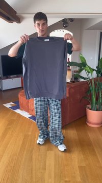 Joven con pantalones de cuadros y zapatillas blancas sosteniendo un suéter gris en una sala de estar moderna con plantas y un televisor.