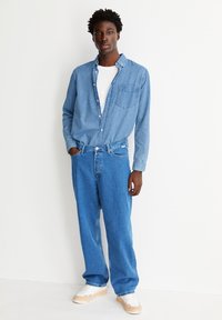 Lichtblauw chambray overhemd met lange mouwen en een voorzak, gecombineerd met loszittende blauwe denim jeans en witte sneakers.