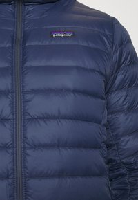 Tamno plava jakna od puf materijala s kvadratnim uzorkom, s prednjim zatvaračem i logom Patagonije na prsima. Mekani, lagani materijal.