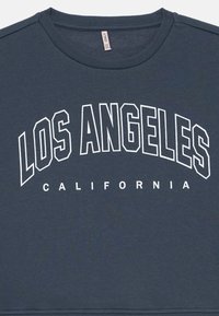 Sötétkék pulóver, amelyen fehér szöveg olvasható: "LOS ANGELES KALIFORNIA" vastag, ívelt betűkkel. Sima textúra, kerek nyakú dizájn.