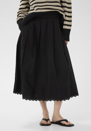 Kobieta ubrana w czarną midi spódnicę z falbankowanym dołem, paskowany sweter z dzianiny oraz czarne sandały z kwadratowym noskiem na gołym szarym tle.