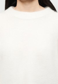 Γυναίκα που φοράει μια μαλακή λευκή μπλούζα με στρόγγυλο λαιμό με μακριά σκούρα μαλλιά, σε απλό φόντο.