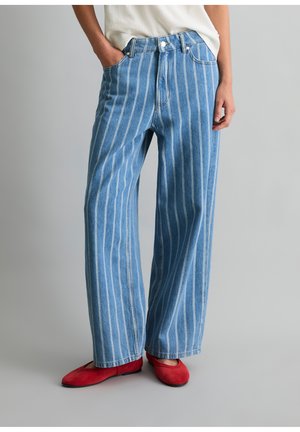 Persoon die blauwe spijkerbroek draagt met witte verticale strepen, rode ballerina's en een wit topje, staand tegen een effen grijze achtergrond.