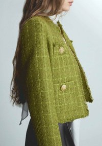 Chaqueta de tweed verde con una forma cuadrada, bordes deshilachados y tejido texturizado. Presenta dos botones en tono dorado y un detalle de bolsillo lateral.