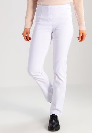 Personne portant un pantalon blanc ajusté, des chaussures noires et un haut beige à manches longues, debout avec un genou légèrement plié.