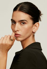 Anillo de oro con un diseño circular en el dedo de un modelo, que lleva pendientes pequeños de aro; el modelo tiene un peinado recogido y viste una blusa negra.