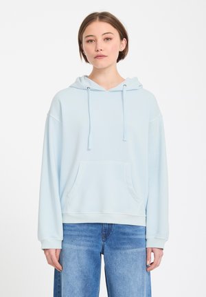 Jeune femme portant un sweat à capuche oversize bleu clair et un jean bleu, debout devant un fond blanc uni.