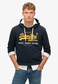 Marineblaue Hoodie mit einer Vordertasche, weißen Kordeln und gelbem Grafiktext "Vintage SuperDry". Getragen über einem kragenlosen weißen Hemd.