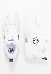 Sapatilhas brancas desportivas com um cabedal sintético texturizado, apresentando um logótipo "N" em azul-marinho, design com atacadores e um colarinho acolchoado para conforto.