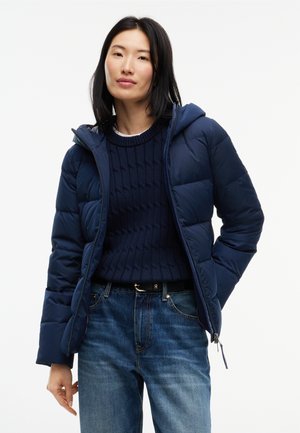 Vrouw draagt een marineblauwe donsjas over een donkerblauwe kabeltrui en blauwe spijkerbroek met een zwarte riem, staand tegen een witte achtergrond.