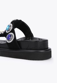 Sandal nero con soletta piatta e cinturino imbottito. Accenti decorativi comprendono strass blu e viola incastonati in minuteria di metallo argentato a forma circolare.