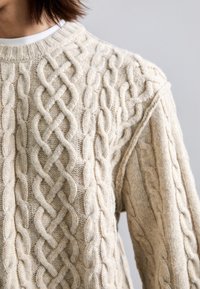 Kreemivärvi nöörkootud kampsun, millel on keerukad punutud mustrid rinnal ja varrukatel, kantud valge särgi peal.