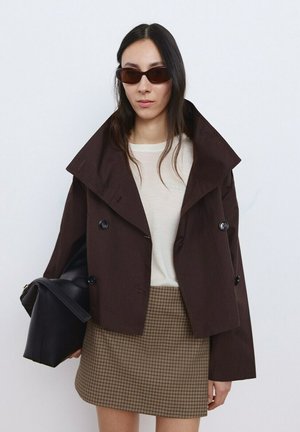 Donna che indossa una giacca corta marrone scuro, top bianco, minigonna a quadri, occhiali da sole, con una grande borsa nera in mano su uno sfondo bianco semplice.