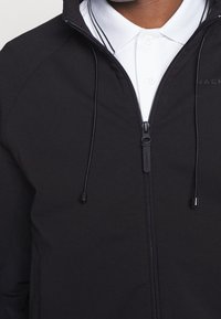 Giacca nera con zip, caratterizzata da un colletto rialzato, coulisse regolabile e tessuto liscio, indossata sopra una camicia bianca con colletto.