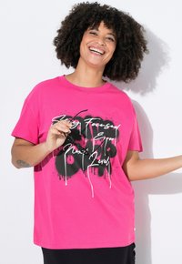 Pink cotton T-shirt with a relaxed fit, featuring black graffiti-style text. Short sleeves and a crew neck, with a small accent detail at the hem.