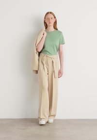T-shirt in cotone verde abbinato a pantaloni beige a gamba larga con vita legata. Il modello tiene in mano un elegante giubbotto beige abbinato. Sneakers bianche completano il look.