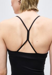 Γυναίκα με κοντά μαλλιά που φοράει μαύρο τοπ με λουράκια, εμφανίζοντας το τριγωνικό σχέδιο στα λουράκια της πλάτης και ασημένια σκουλαρίκια κρίκους.