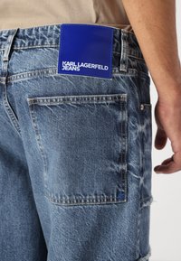 Traperice u svijetloplavoj boji s izblijedjelom teksturom, s plavom etiketom na pojasu na kojoj piše "KARL LAGERFELD JEANS" bijelim slovima.