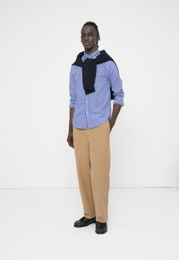 Πουκάμισο μπλε και λευκό καρό, σκούρο μπλε πουλόβερ περασμένο στους ώμους, μπεζ παντελόνι φαρδύ, μαύρα παπούτσια· απλός σχεδιασμός και άνετη εφαρμογή.