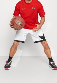 Jugador de baloncesto masculino vestido con una camiseta roja de los Chicago Bulls y pantalones cortos blancos de Nike, driblando un balón de baloncesto Spalding en una postura atlética.