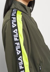 Zielona kurtka zapinana na zamek z wysokim kołnierzem, z czarno-białą taśmą z logo FILA oraz jasnoniebieskim paskiem po boku. Gładka tekstura materiału.