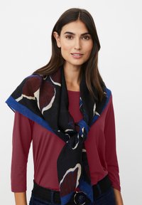 Mujer con cabello largo y oscuro, vestida con una blusa color burdeos y una bufanda negra con patrones de hojas azules y marrones atada alrededor de su cuello.