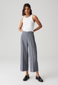 Pantalons larges en tissu texturé bleu marine avec de légers éclats. Taille haute avec plis à l'avant. Associés à un haut sans manches blanc et des mocassins noirs.